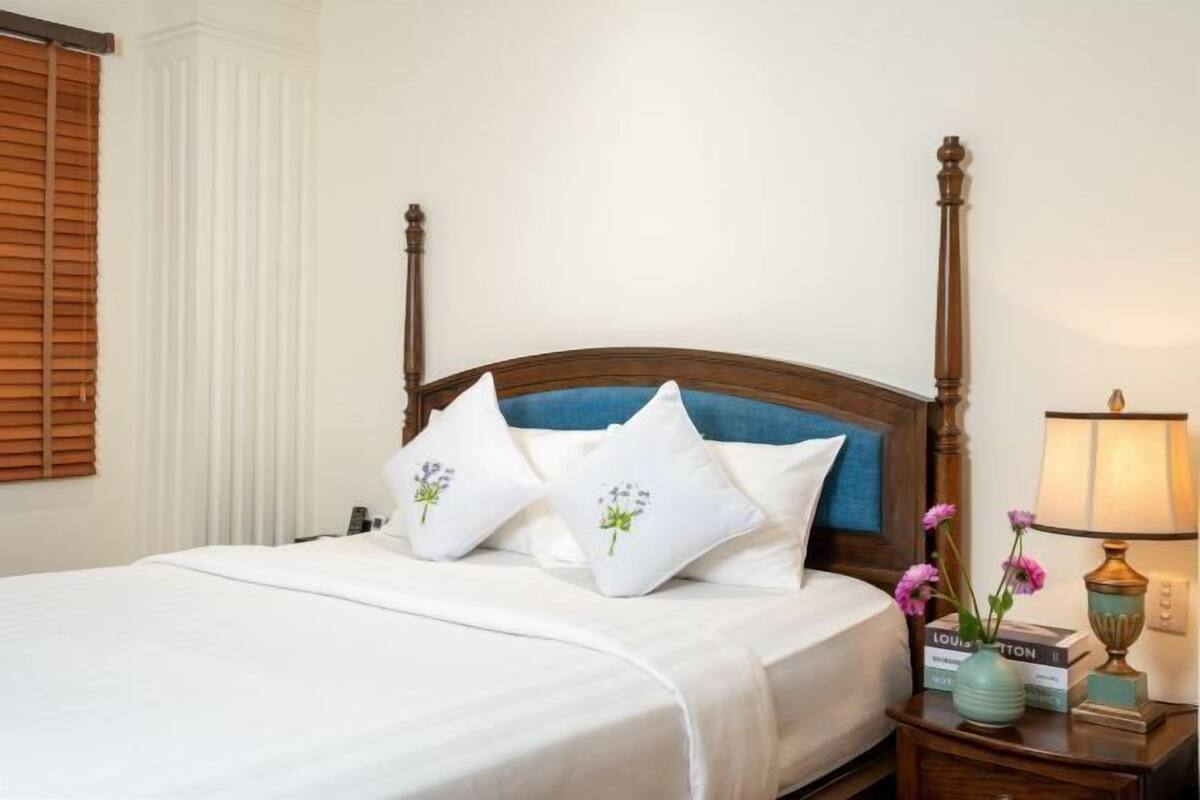 A neatly made bed with white bedding and decorative pillows, a wooden headboard, a bedside table with a lamp, and a book.