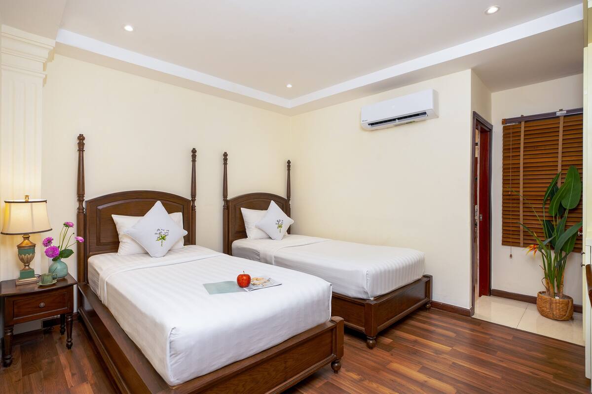 A hotel room with two wooden beds, a nightstand with a lamp, a vase with flowers, and a potted plant.