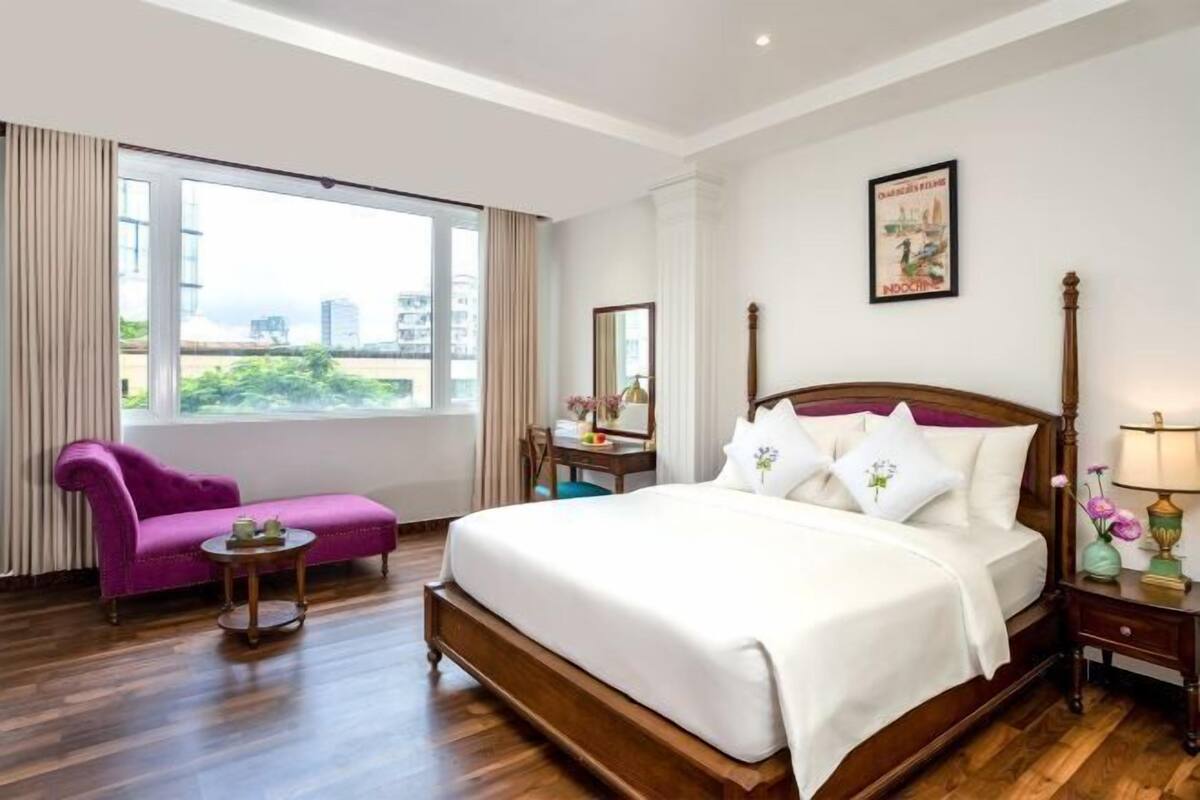 A hotel room with a large bed, a purple armchair, a small table, a nightstand, and a window with a city view.
