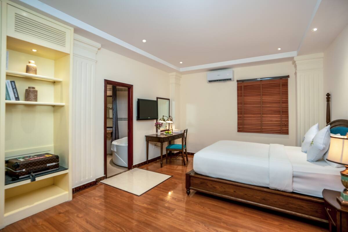 A hotel room with a wooden floor, a bed, a desk with a chair, a wardrobe, and a television.