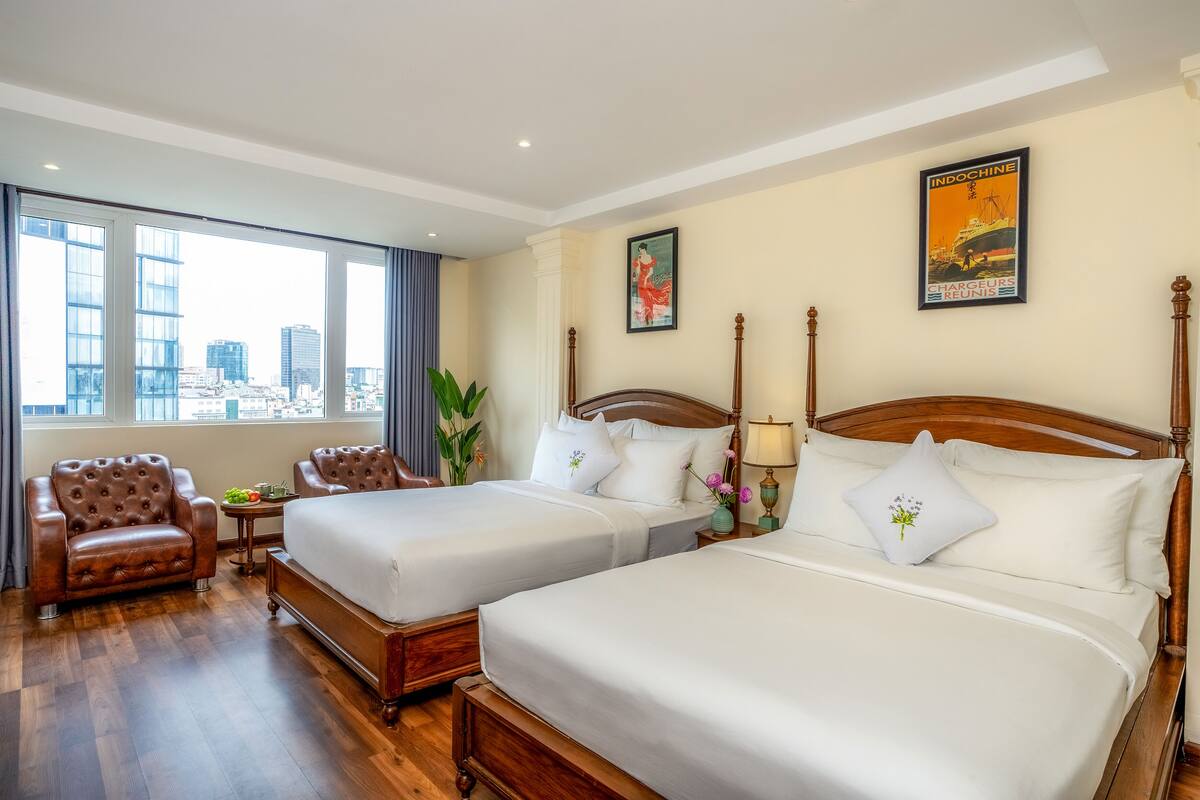 A hotel room with two beds, a leather armchair, a small table with a plant, and a city view.