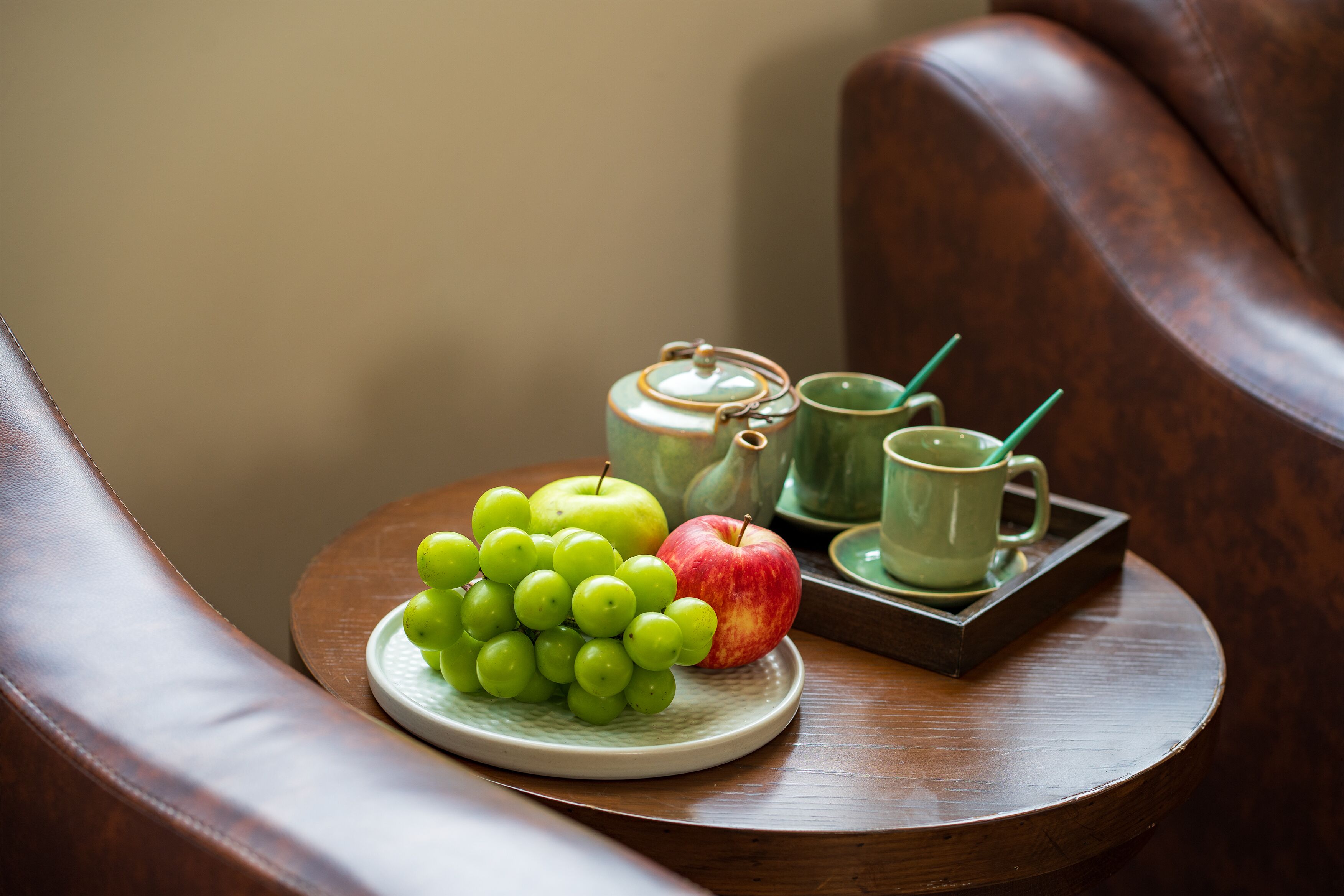A wooden table with a plate of green grapes and an apple, a green cup, and a teapot.