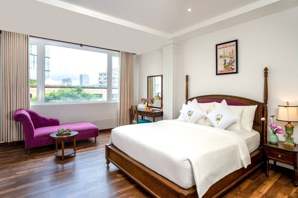 A hotel room with a large bed, a purple armchair, a small table, a nightstand with a lamp, and a window with curtains.