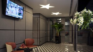 Um saguão com piso xadrez, duas cadeiras vermelhas, uma mesa com frutas, uma TV exibindo um noticiário e uma grande planta em vaso.