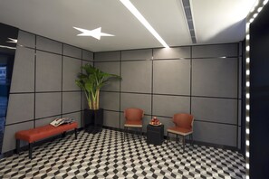 A modern waiting area with a checkered floor, a bench, two chairs, a small table with fruit, and a potted plant. - Hotel 81 Premier Hollywood (Singapore)