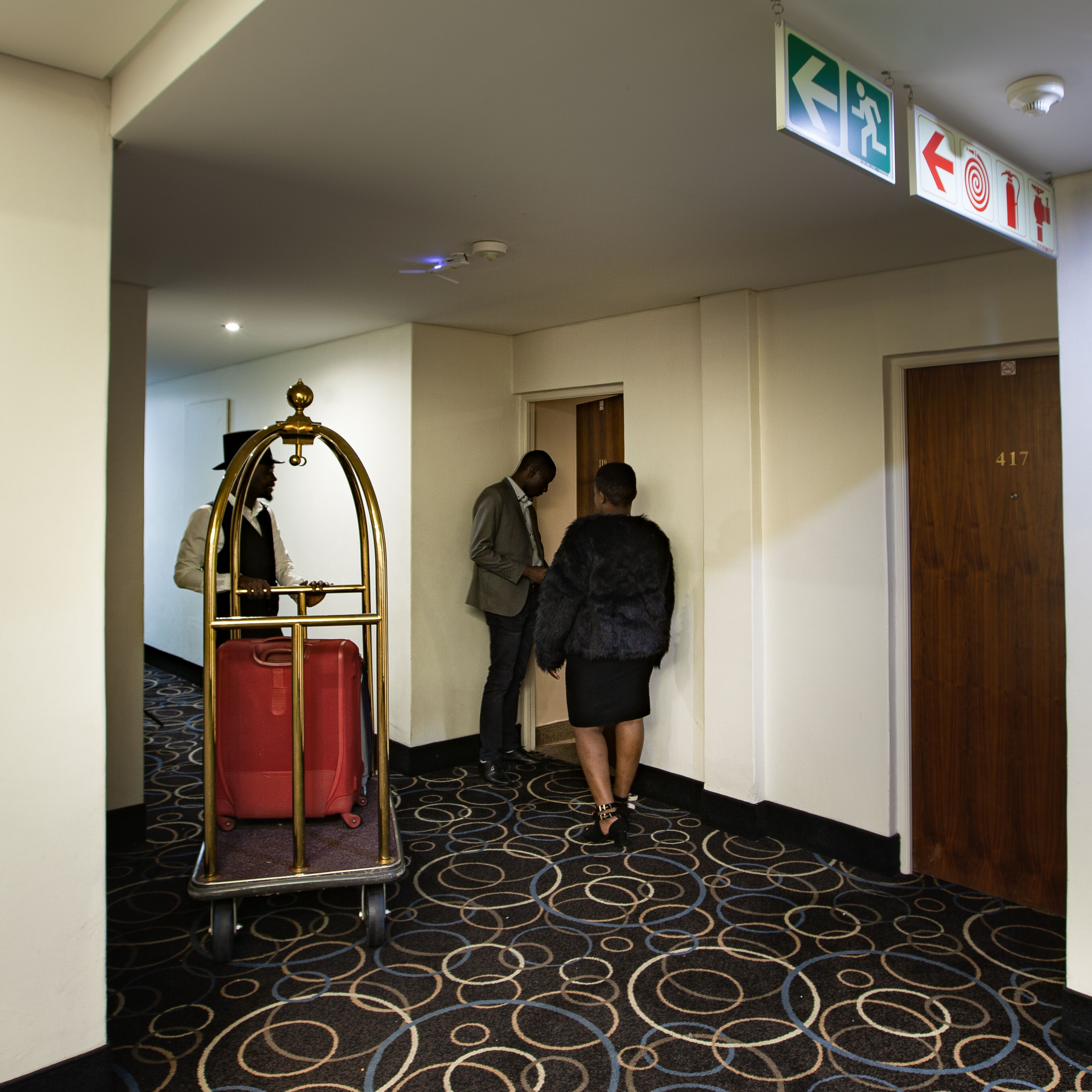 A hotel hallway with a patterned carpet, a red suitcase on a trolley, and a wooden door marked with the number 417.