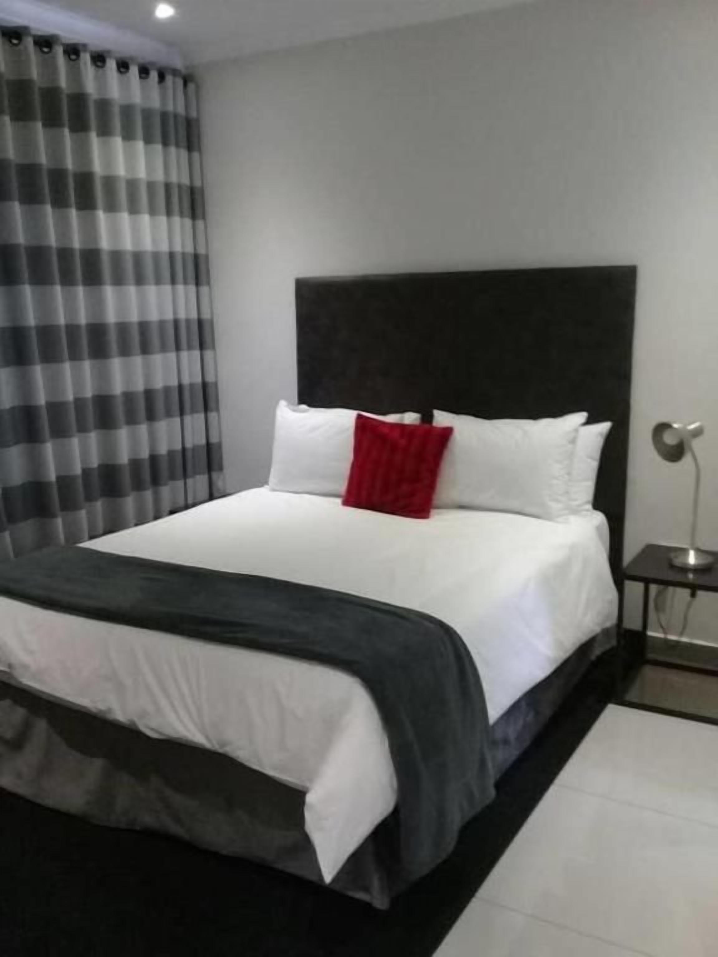 A hotel room with a large bed, a black headboard, white bedding, a red pillow, a small bedside table, and a curtain.