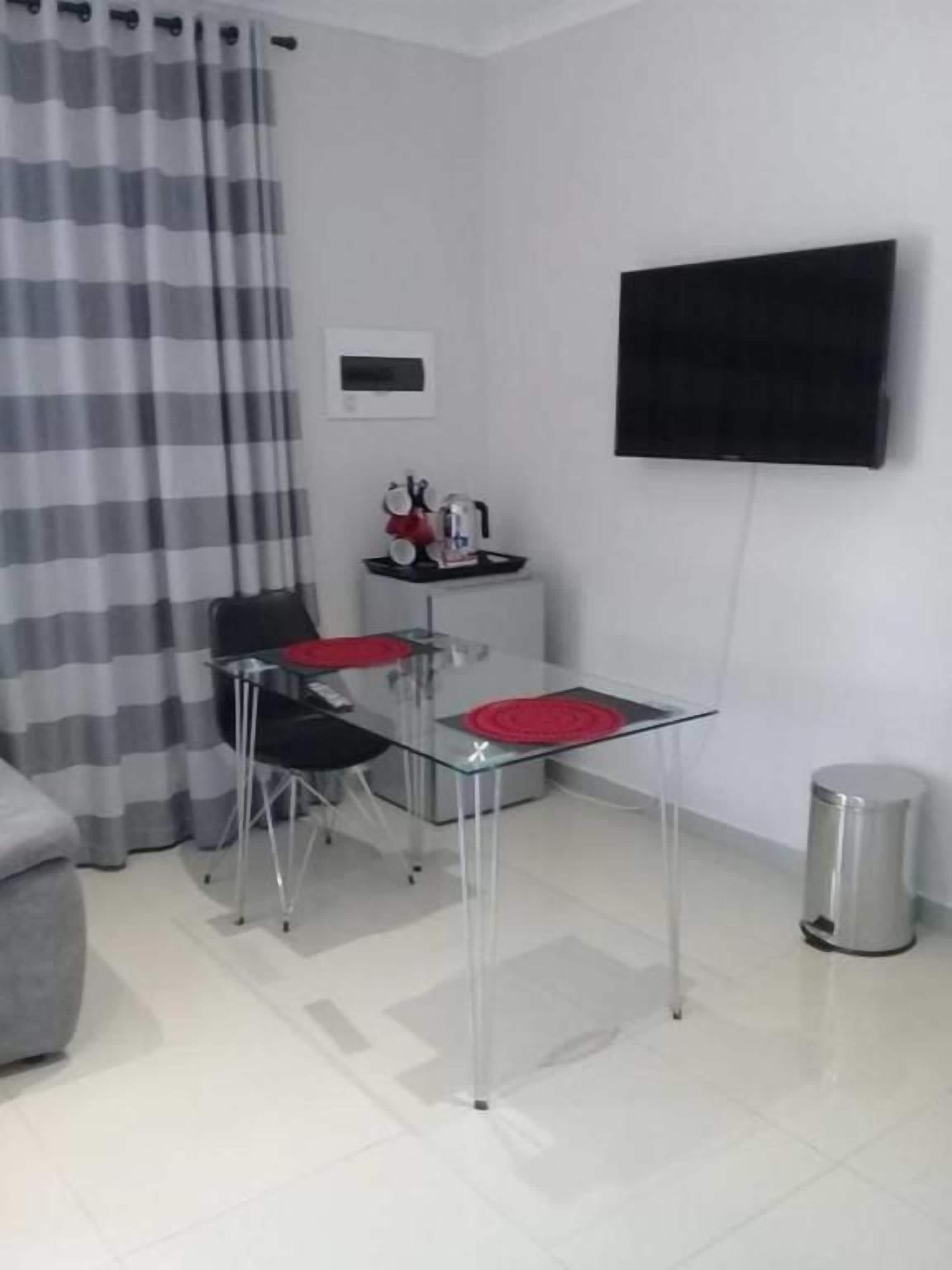 Une petite pièce moderne comprenant une table à manger, deux chaises, un canapé gris, un téléviseur fixé au mur et une poubelle.