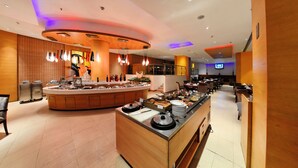 Un comedor de hotel moderno con un bufé, una pantalla de televisión y un área de estar con mesas y sillas.