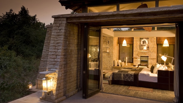 A modern bedroom with a stone fireplace, a large bed, and a glass door leading to a balcony. - Pashan Garh, Panna National Park (Panna)
