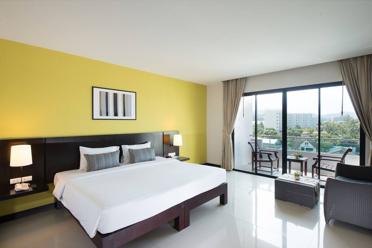 Habitación de hotel con una cama grande, lámparas de noche, una mesita, una silla y un balcón con vistas a edificios.