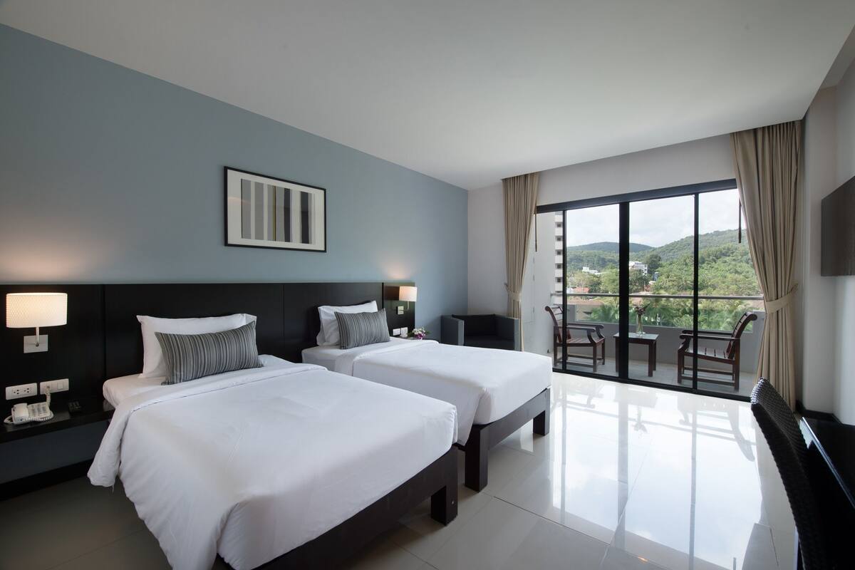 Habitación de hotel con dos camas, un ventanal con vistas a zonas verdes, un televisor de pantalla plana y un balcón con mobiliario.