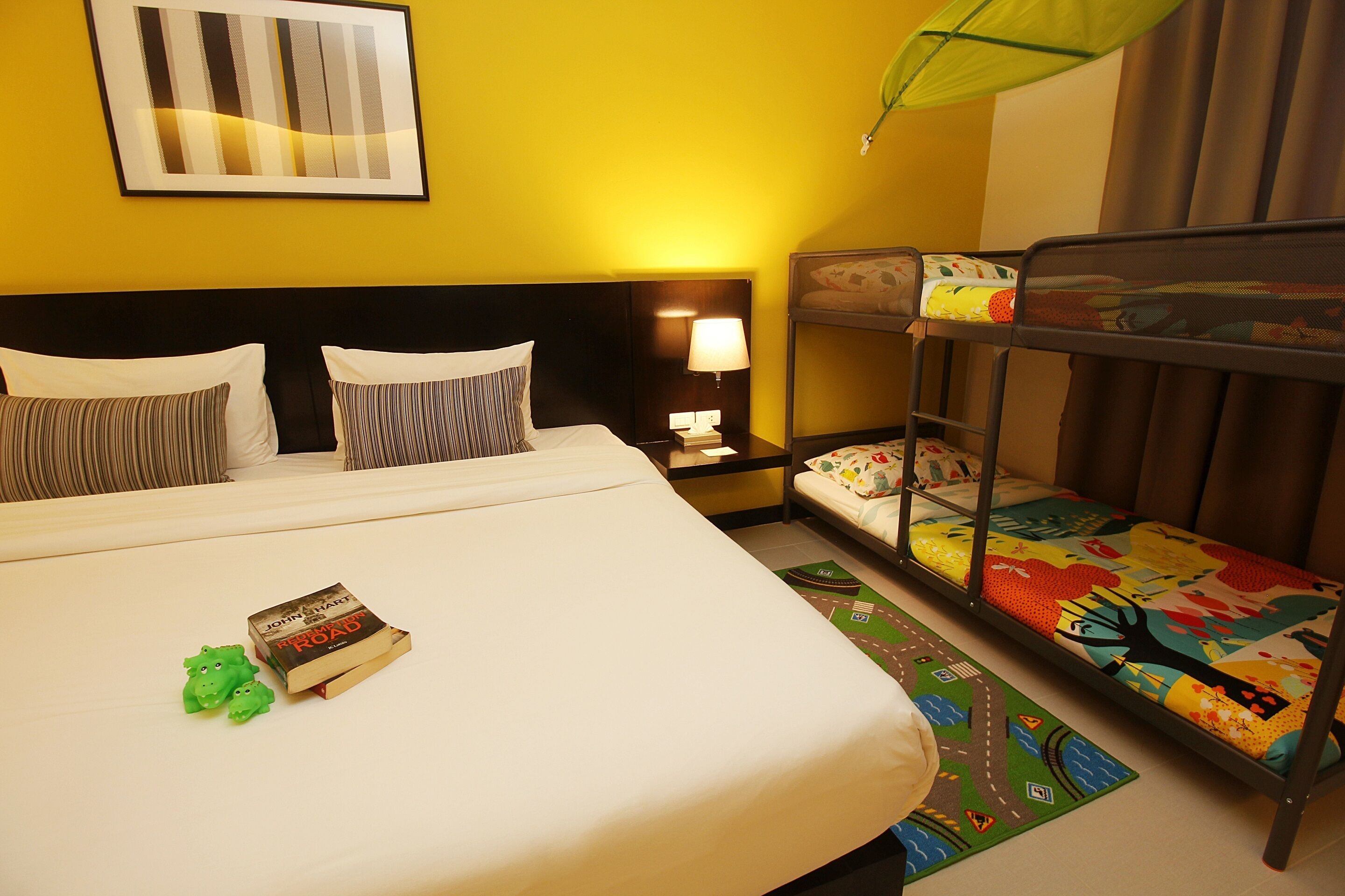 A hotel room with a large bed, bunk beds, a bedside lamp, a framed picture on the wall, and a green umbrella.