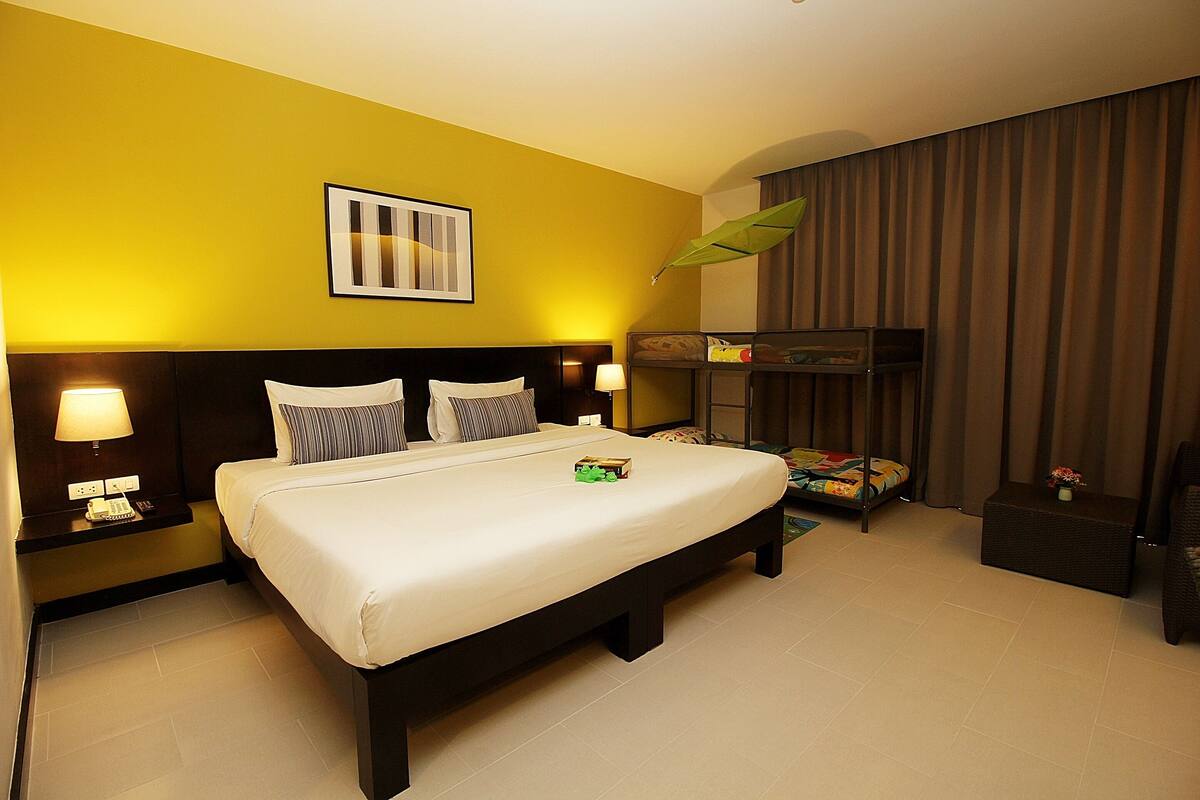 Una habitación de hotel con una cama grande, mesitas de noche con lámparas, una pequeña mesita auxiliar con un jarrón y un ventanal con cortinas.