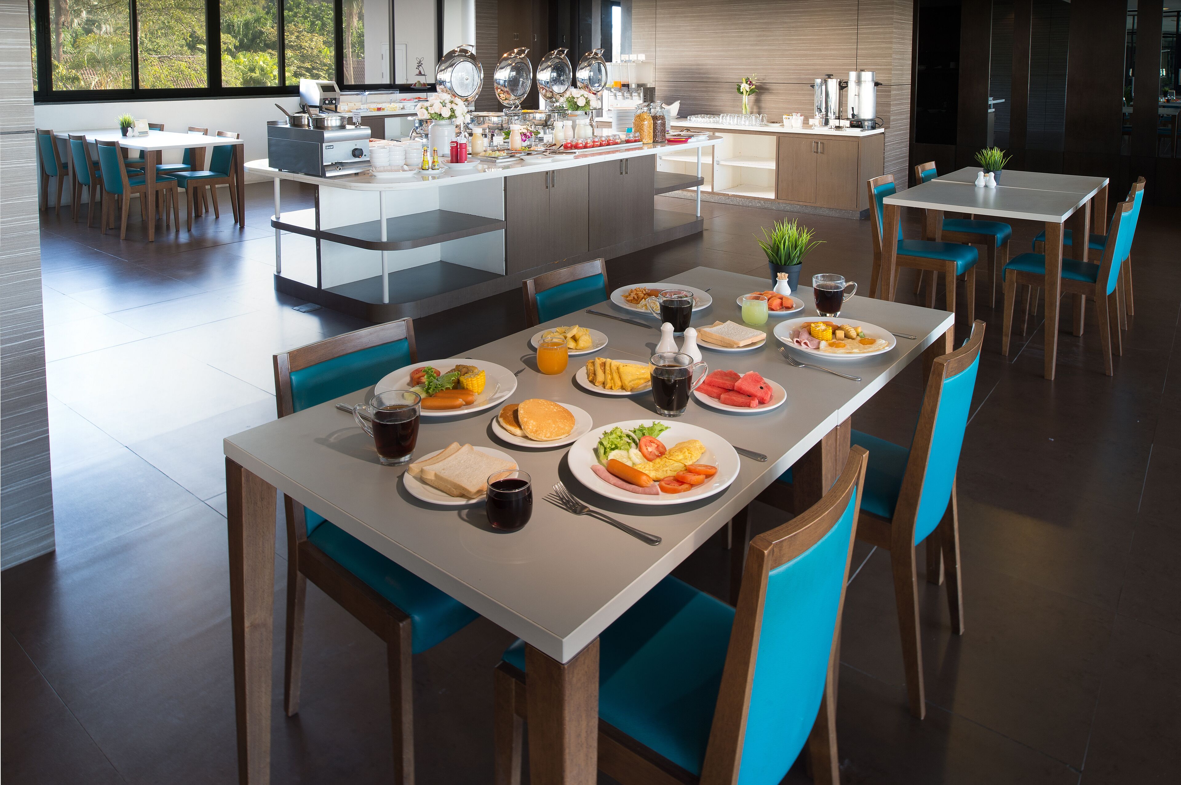 A modern dining area with multiple tables and chairs, a kitchen area with appliances, and a view of greenery through large windows.