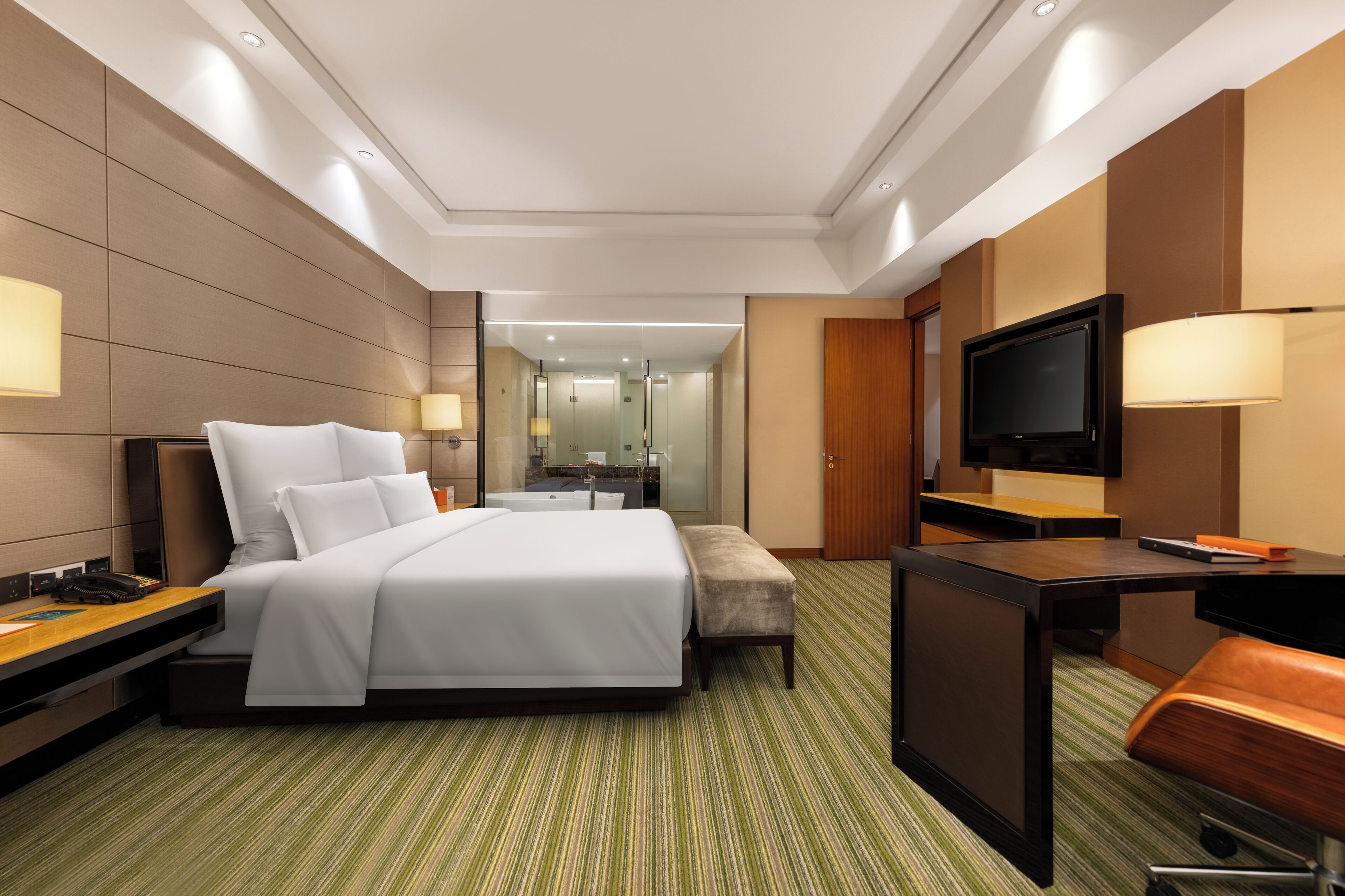 A hotel room with a bed, a desk, a television, and a bathroom visible through a glass door.