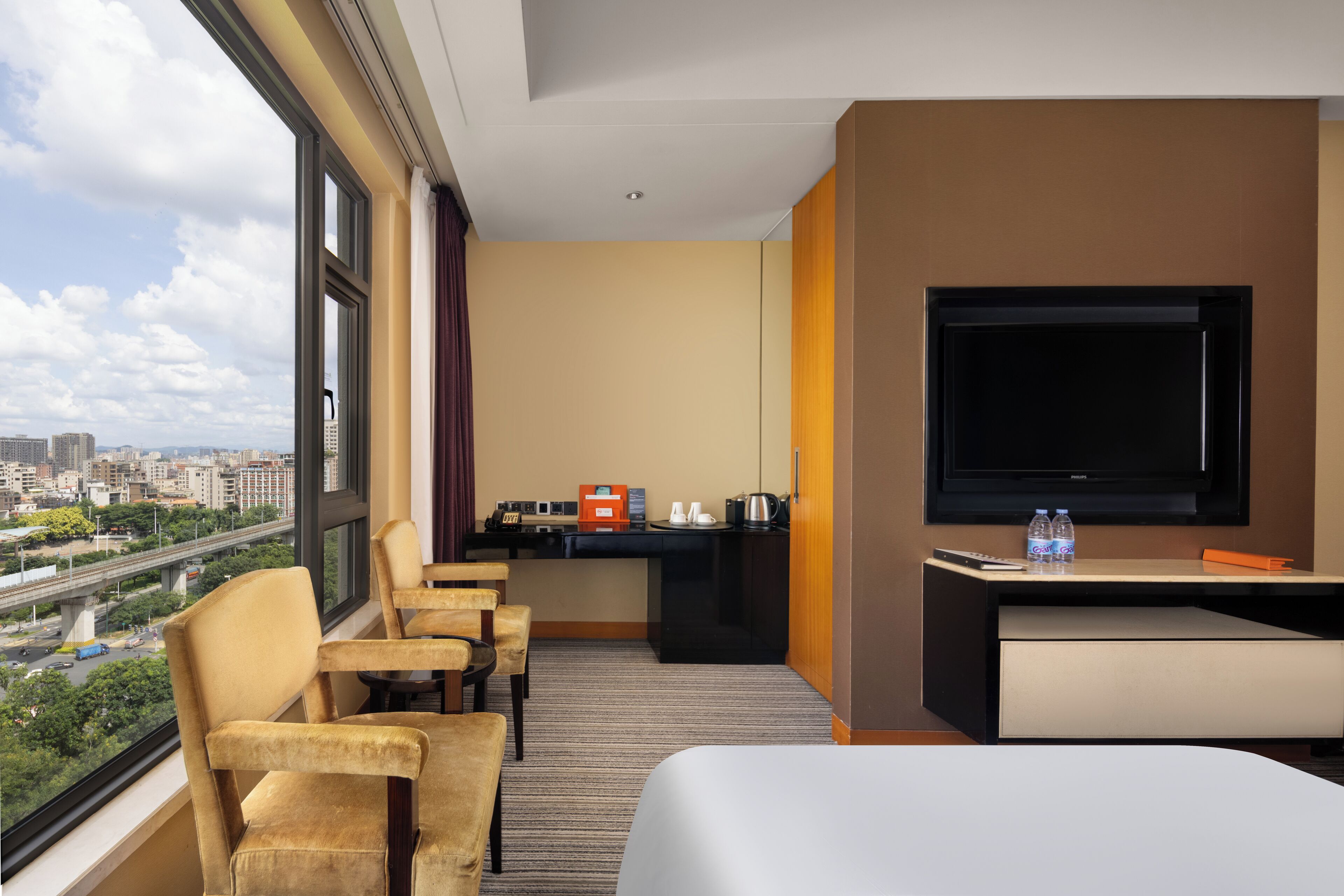 A hotel room with a large window, a bed, a desk with a TV, and a seating area with chairs.