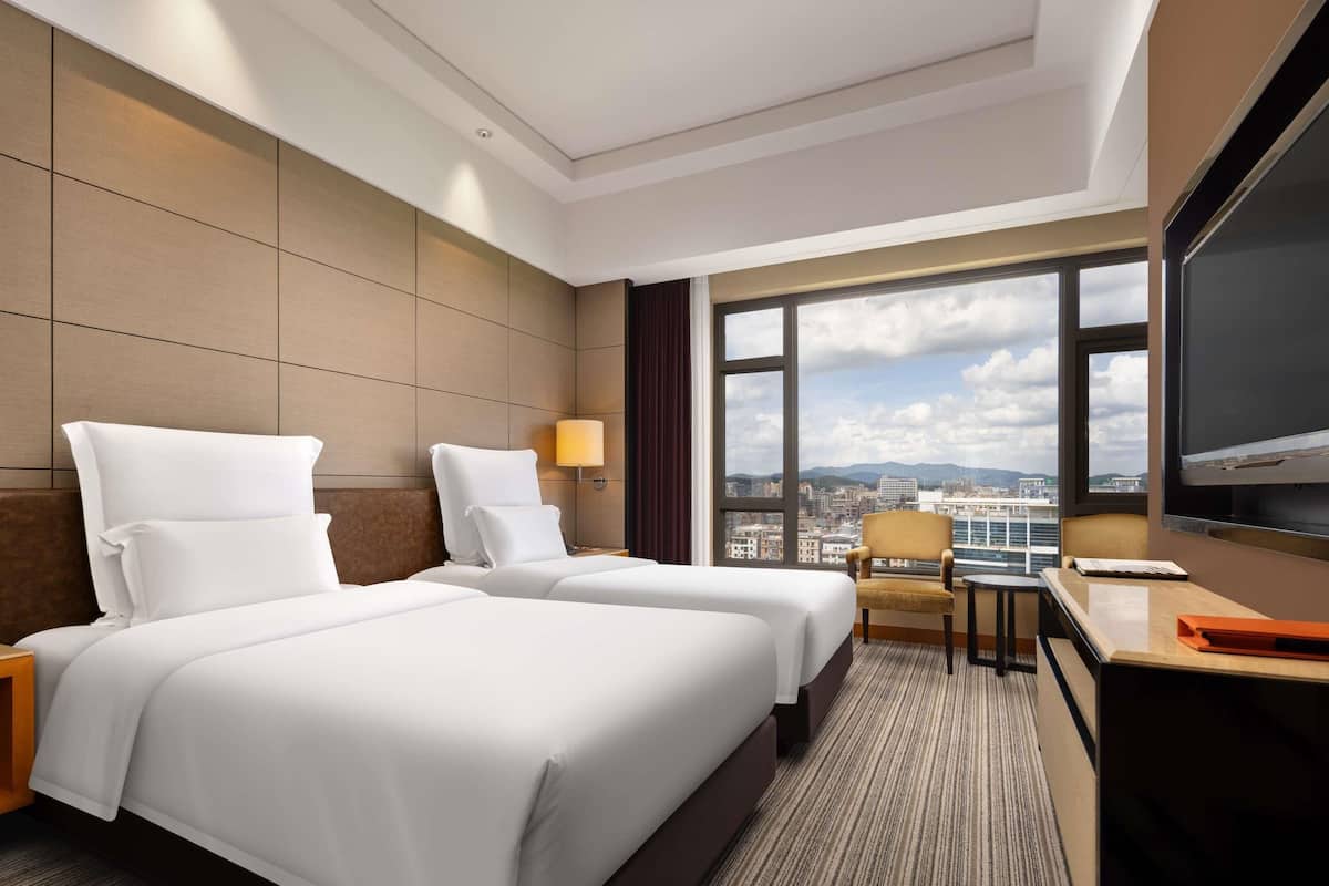 Habitación de hotel con dos camas, un escritorio, una silla, un televisor y un ventanal grande con vistas a la ciudad.