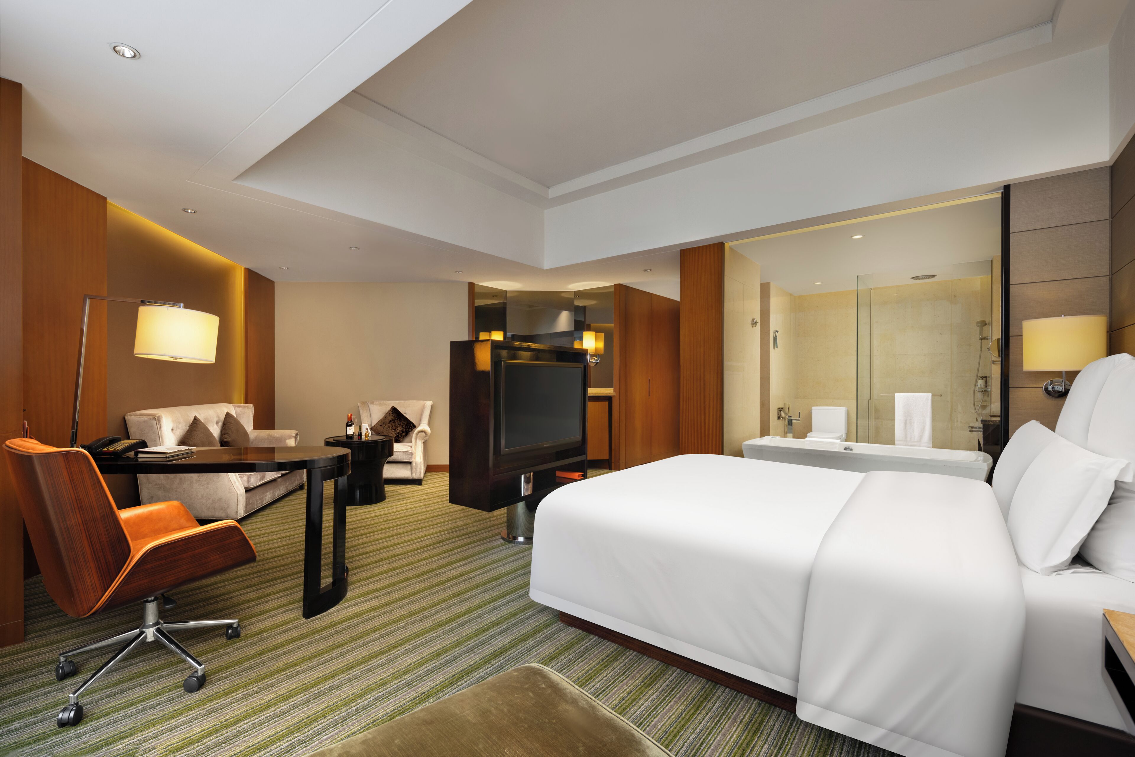 A hotel room with a large bed, a desk with a chair, a television, a sofa, and a bathroom with a mirror and shower.