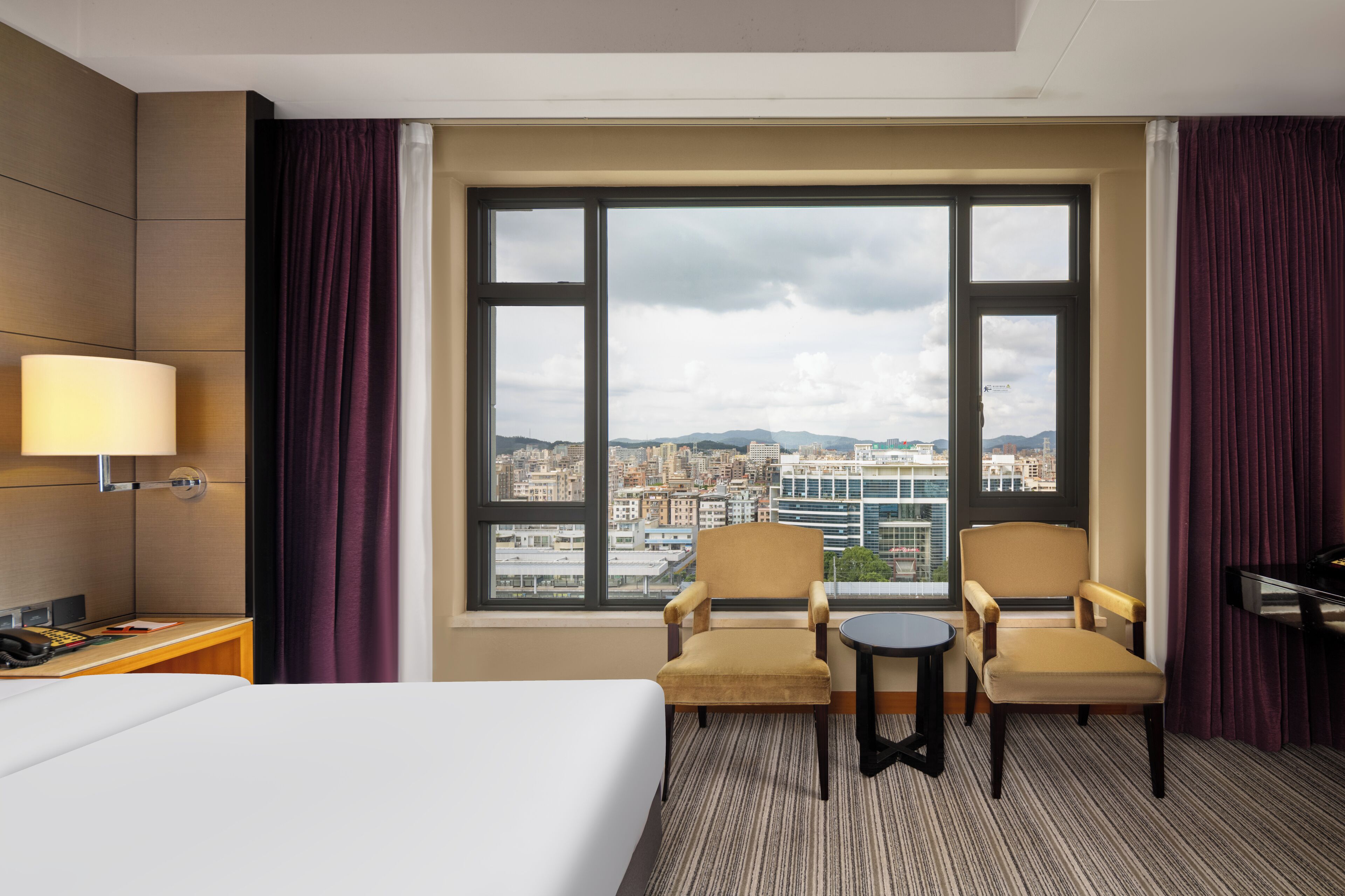 A hotel room with a large window, two armchairs, a small table, a bed, a desk with a lamp, and a piano.