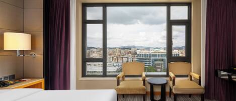 A hotel room with a large window, two armchairs, a small table, a bed, a desk with a lamp, and a piano.
