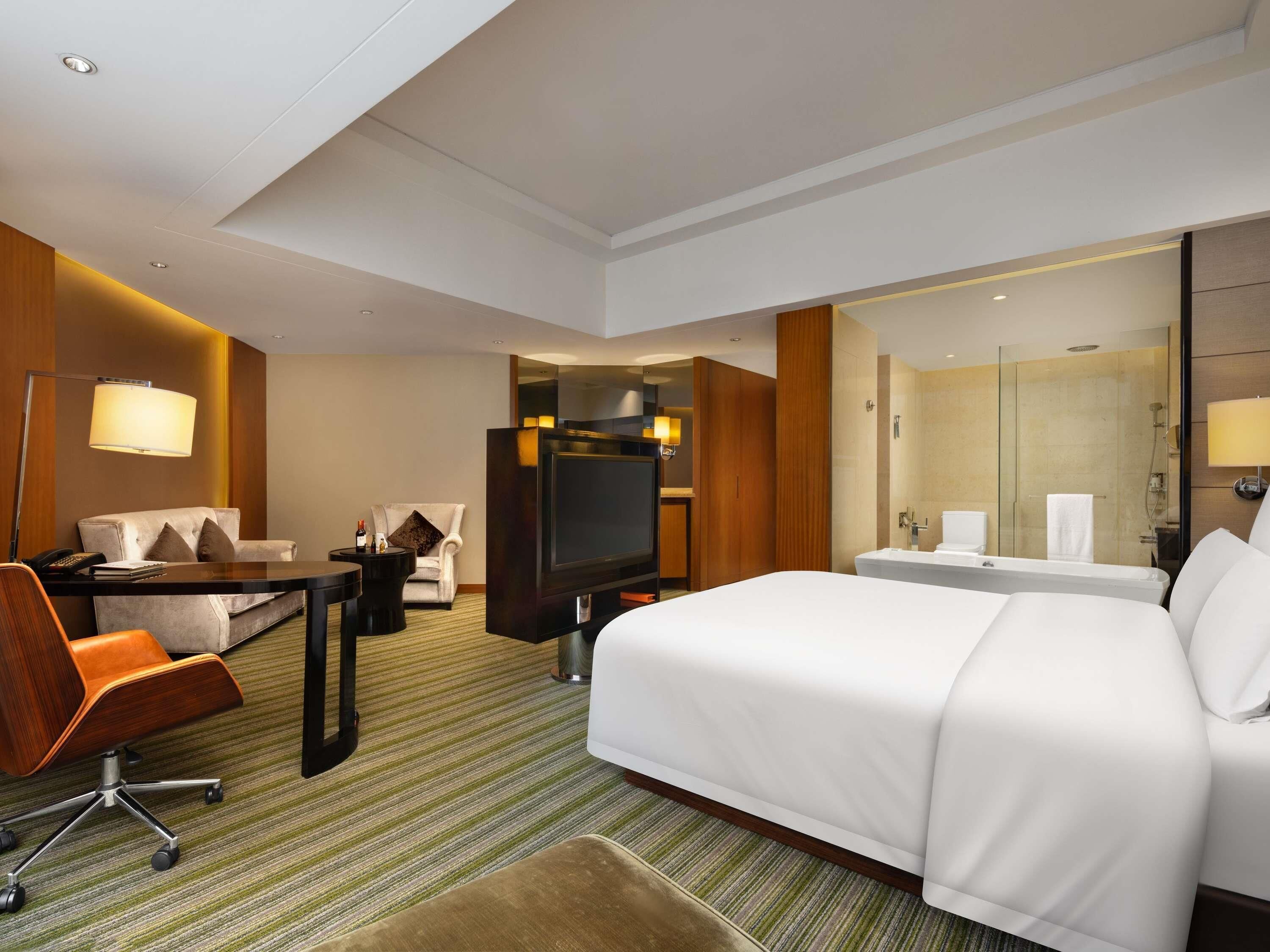 A hotel room with a large bed, a desk with a chair, a TV, a sofa, and a bathroom with a mirror and sink.