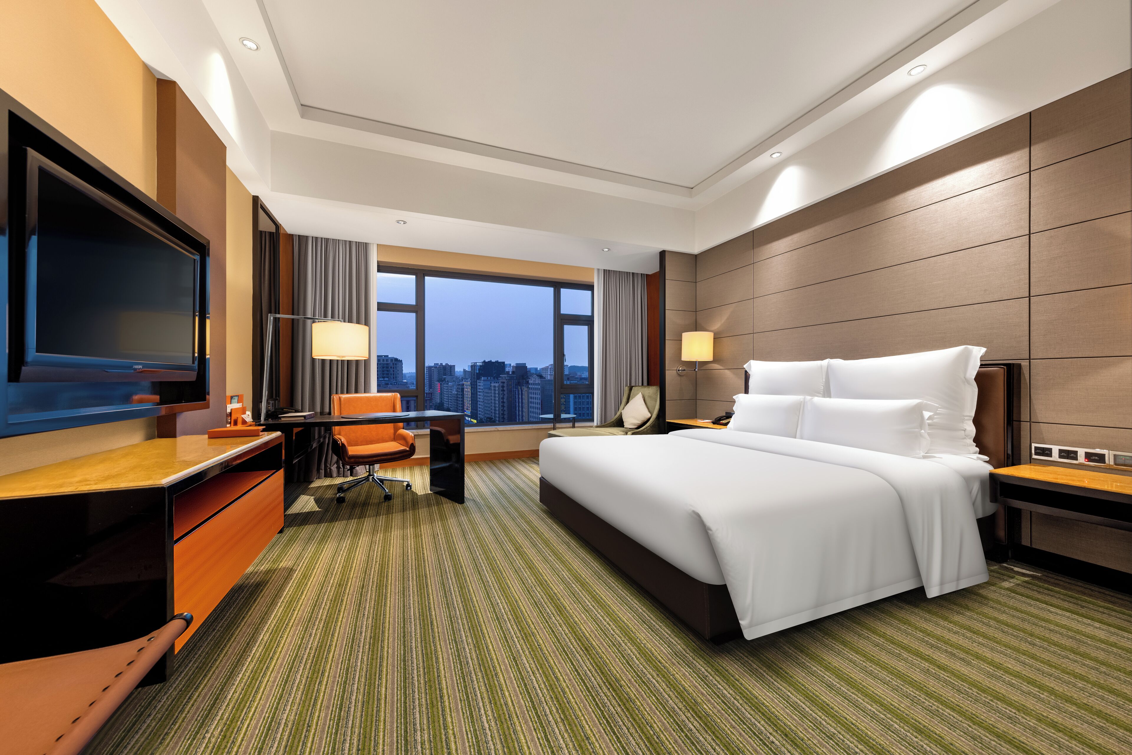 A hotel room with a large bed, a desk with a chair, a television, and a city view.