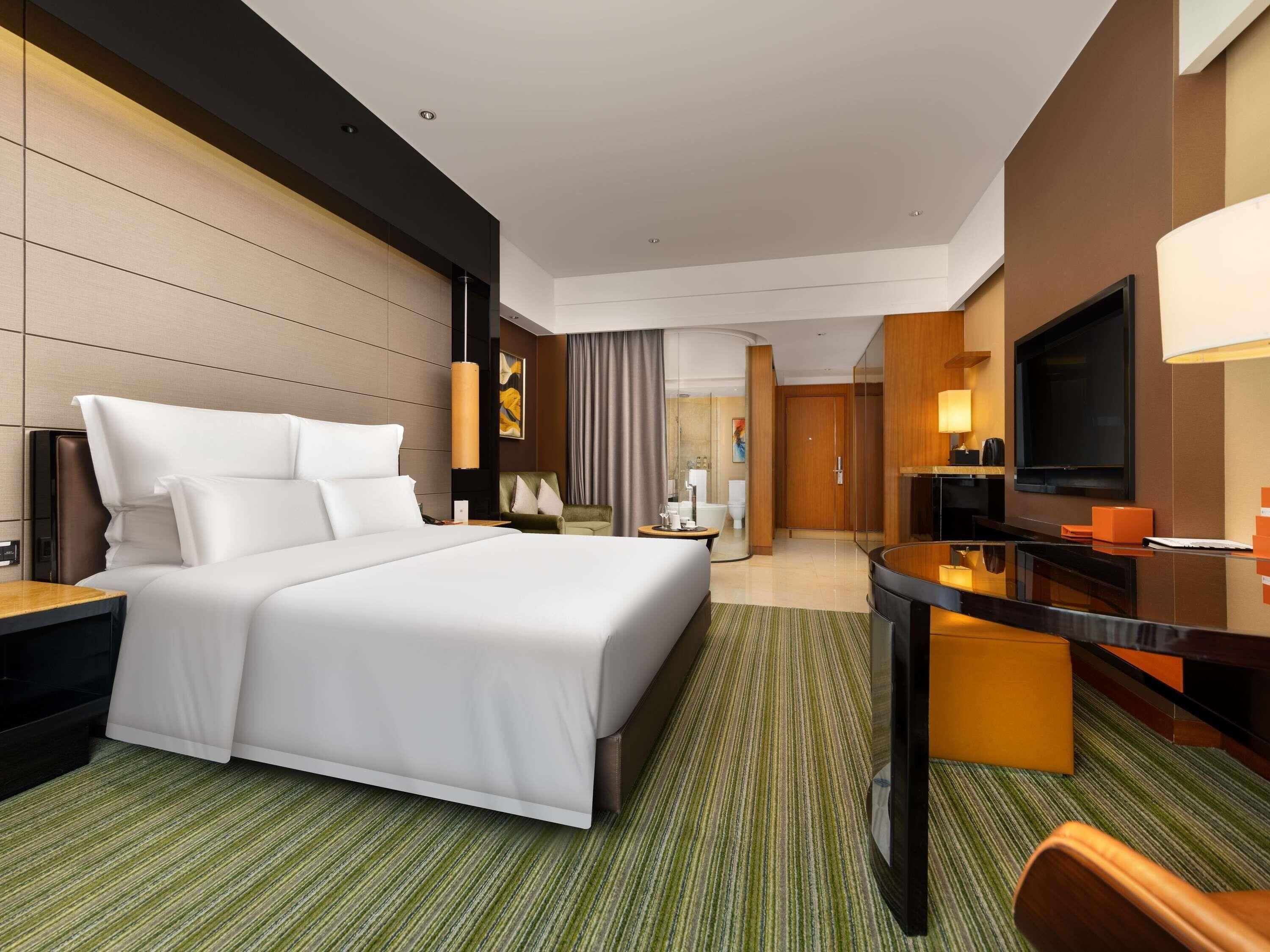 Quarto de hotel moderno com uma cama grande, uma escrivaninha, uma televisão e um banheiro visível através de um painel de vidro.