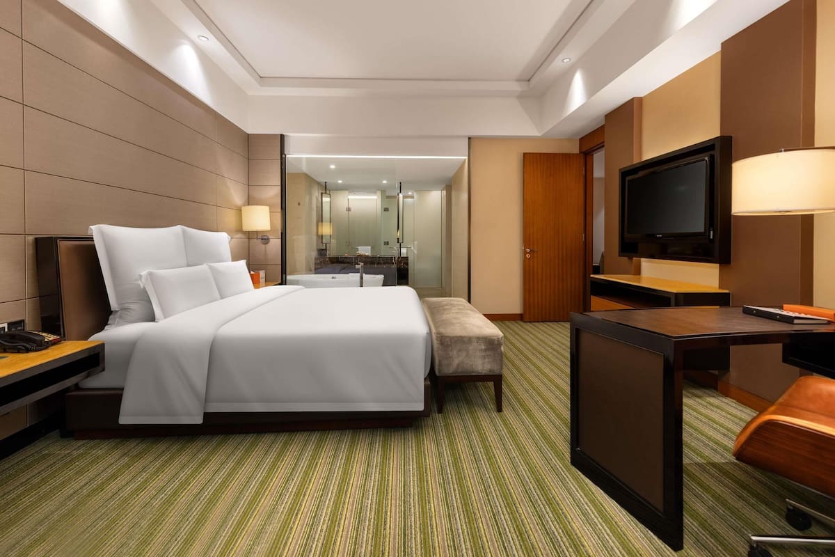 Habitación de hotel con una cama grande, un escritorio con televisor, una silla y un baño visible a través de un panel de vidrio.