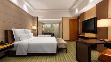 A hotel room with a large bed, a desk with a TV, a chair, and a bathroom visible through a glass partition.