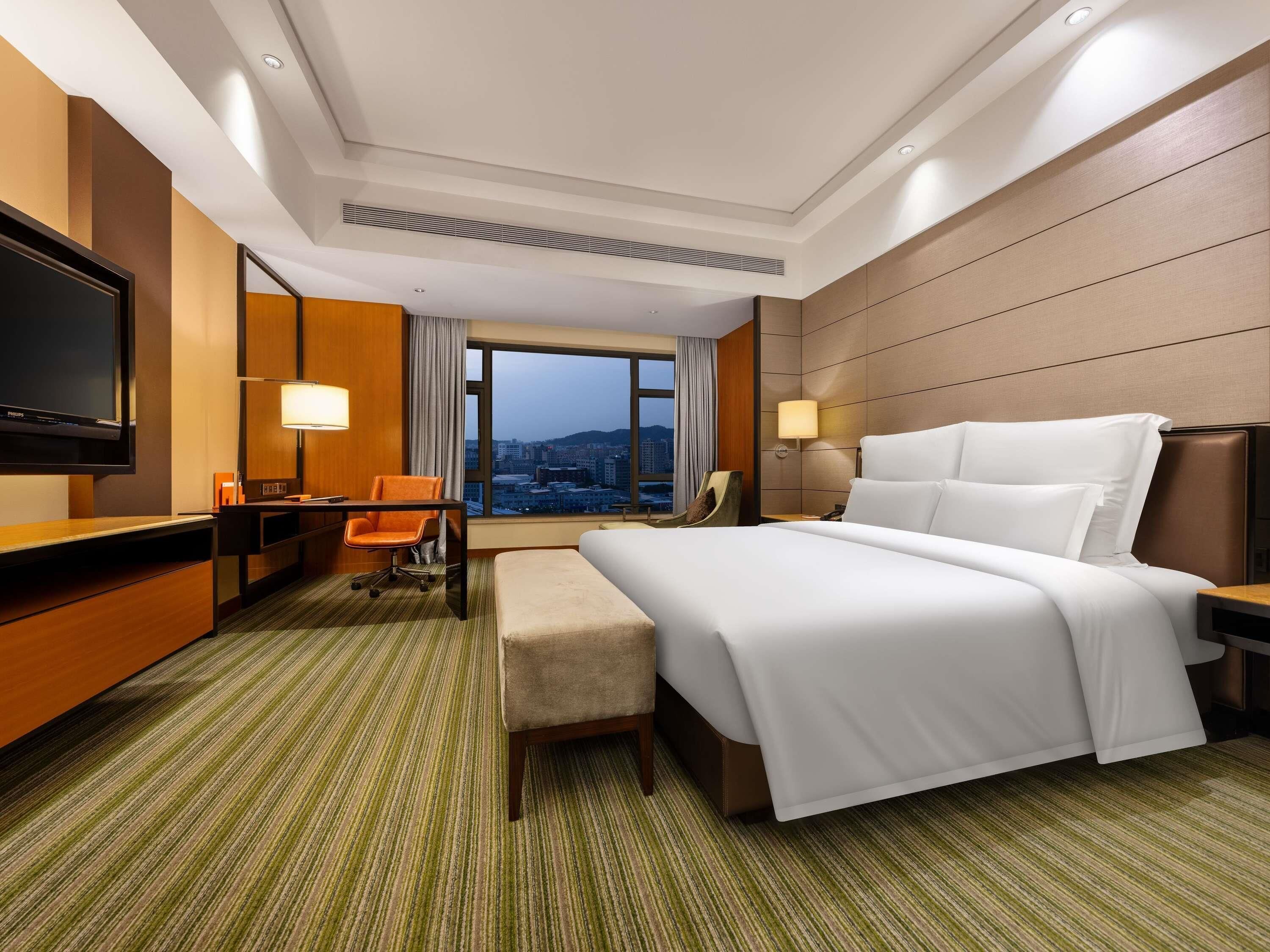 A hotel room with a bed, desk, chair, TV, and a view of the city.