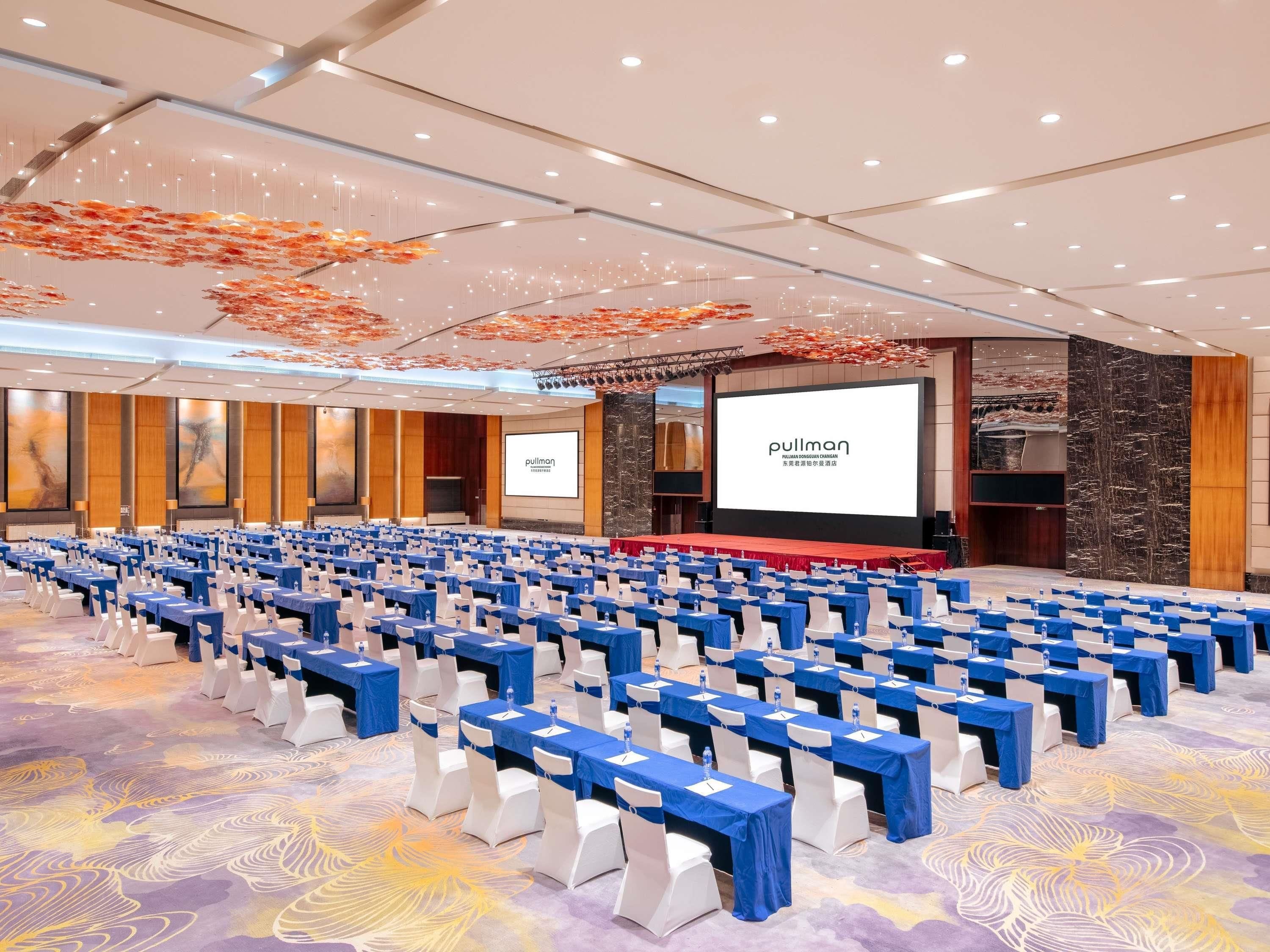 A conference room with rows of tables and chairs, a stage with a screen displaying 'pullman', and a large screen in the background.