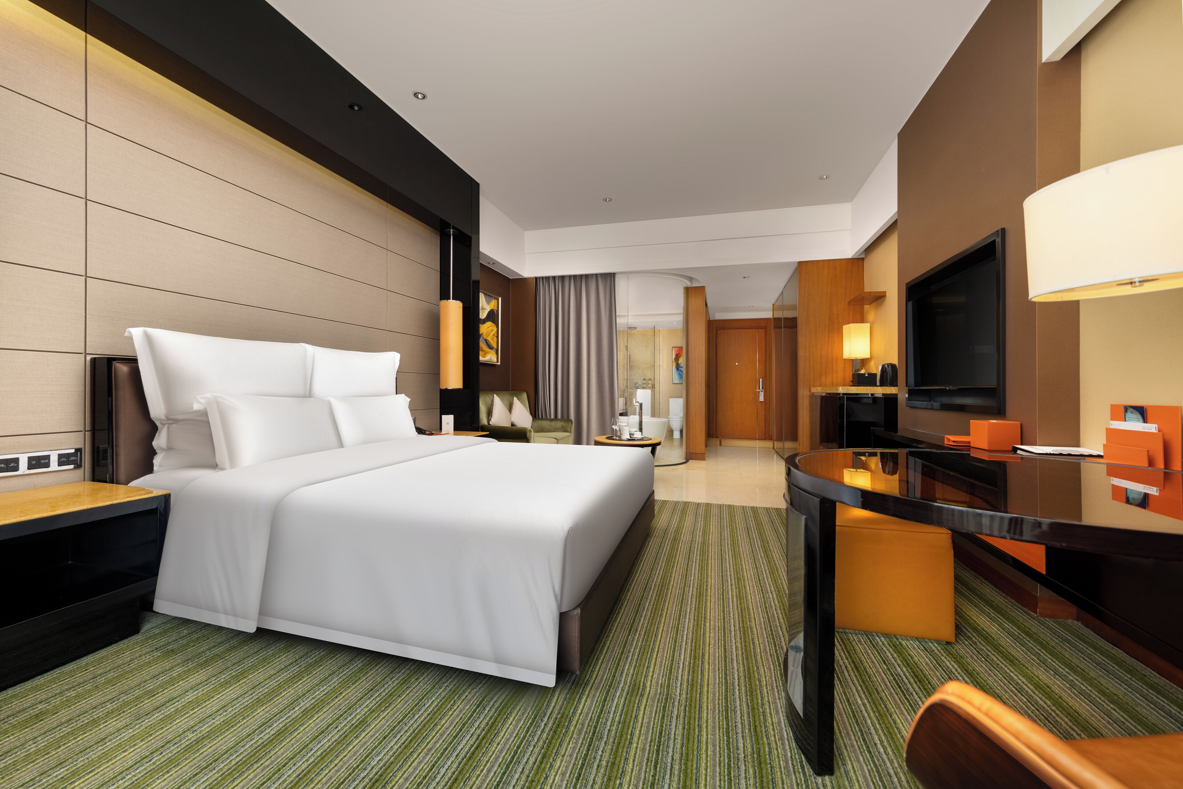 A modern hotel room with a large bed, a desk, a TV, and a bathroom visible in the background.