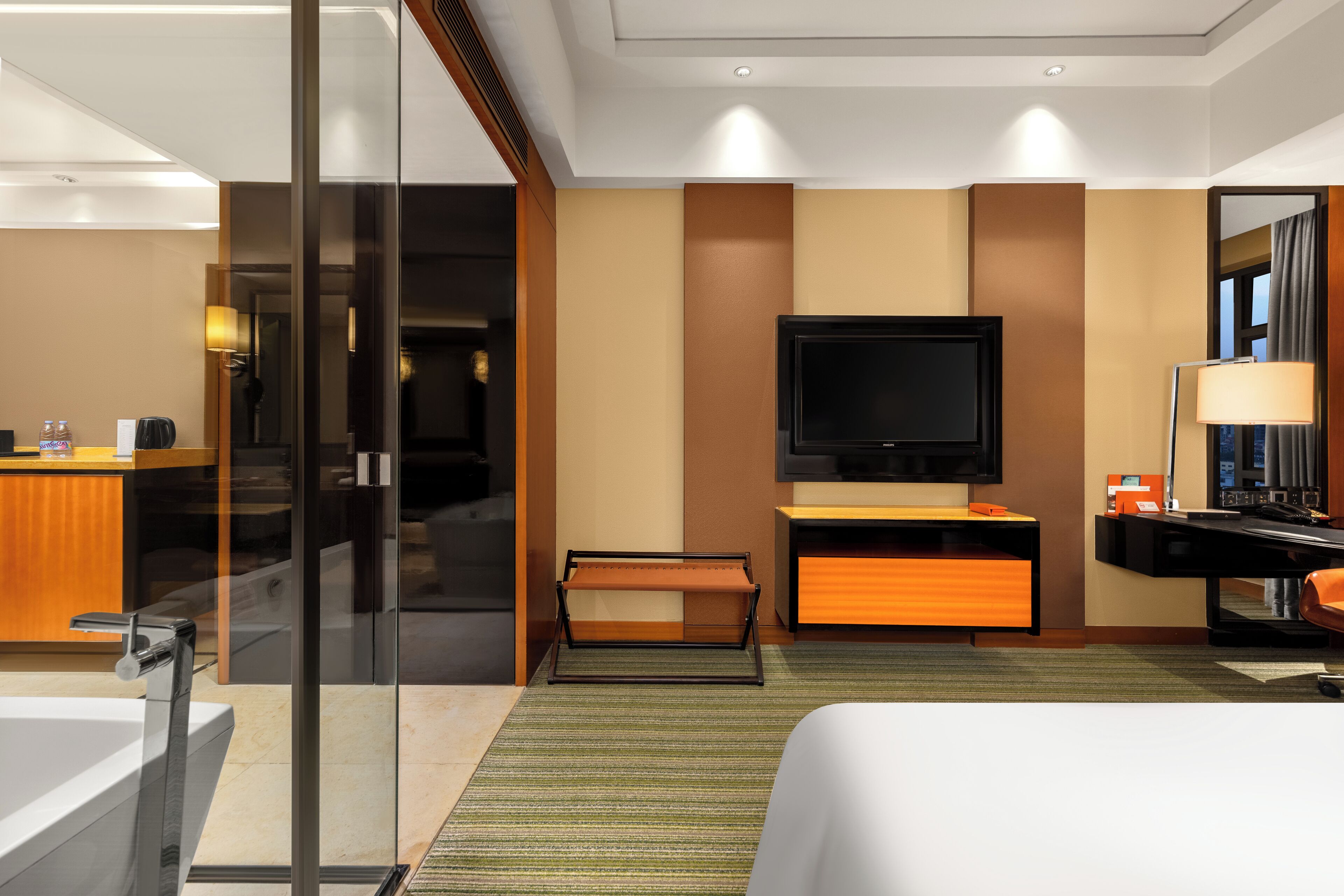 Una habitación de hotel moderna con una cama grande, un televisor de pantalla plana montado en la pared, un escritorio con lámpara y una zona de ducha con cerramiento de vidrio.