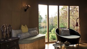 A modern living room with a curved sofa, a wooden bench, and a view of a garden through large windows. - Banjaar Tola, Kanha National Park (Baihar)