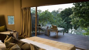 A bedroom with a large bed, a small table, and a view of a wooden deck and a lake. - Banjaar Tola, Kanha National Park (Baihar)