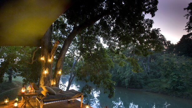 A wooden deck with a bed and lanterns, overlooking a calm river and dense trees. - Banjaar Tola, Kanha National Park (Baihar)