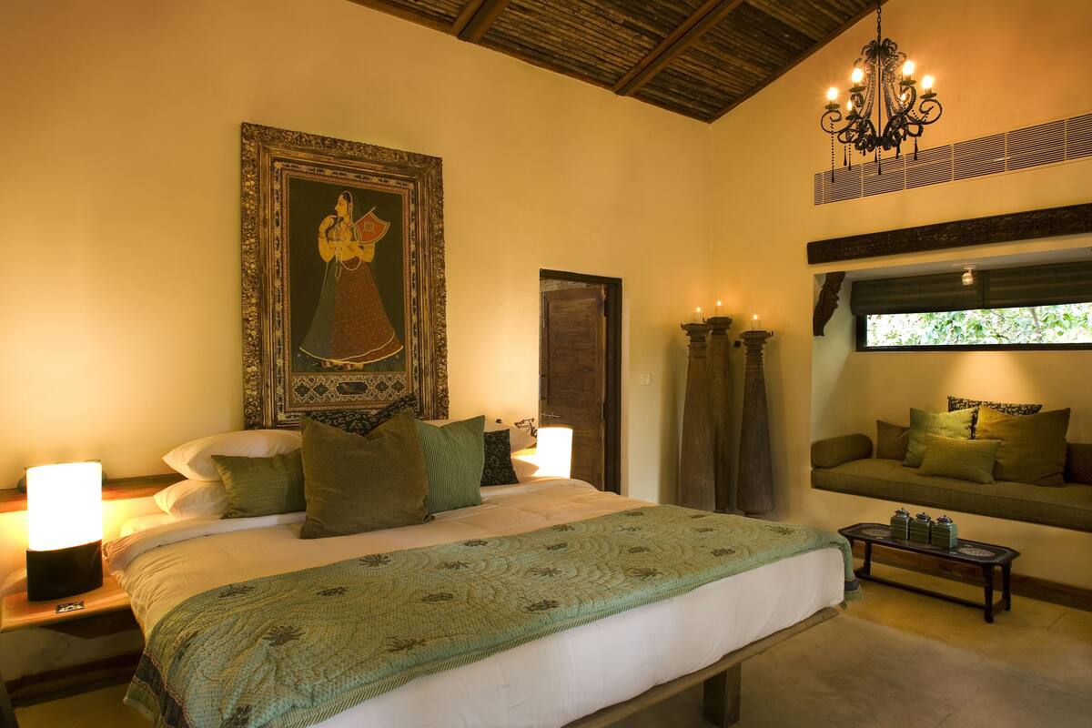 A bedroom with a large bed, a nightstand with a lamp, a window seat with cushions, and a painting on the wall.