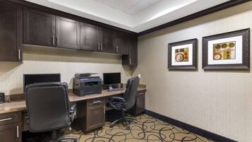 A hotel room with a desk, two monitors, a printer, and a black office chair. There are framed pictures on the wall and a carpet with a swirl pattern.