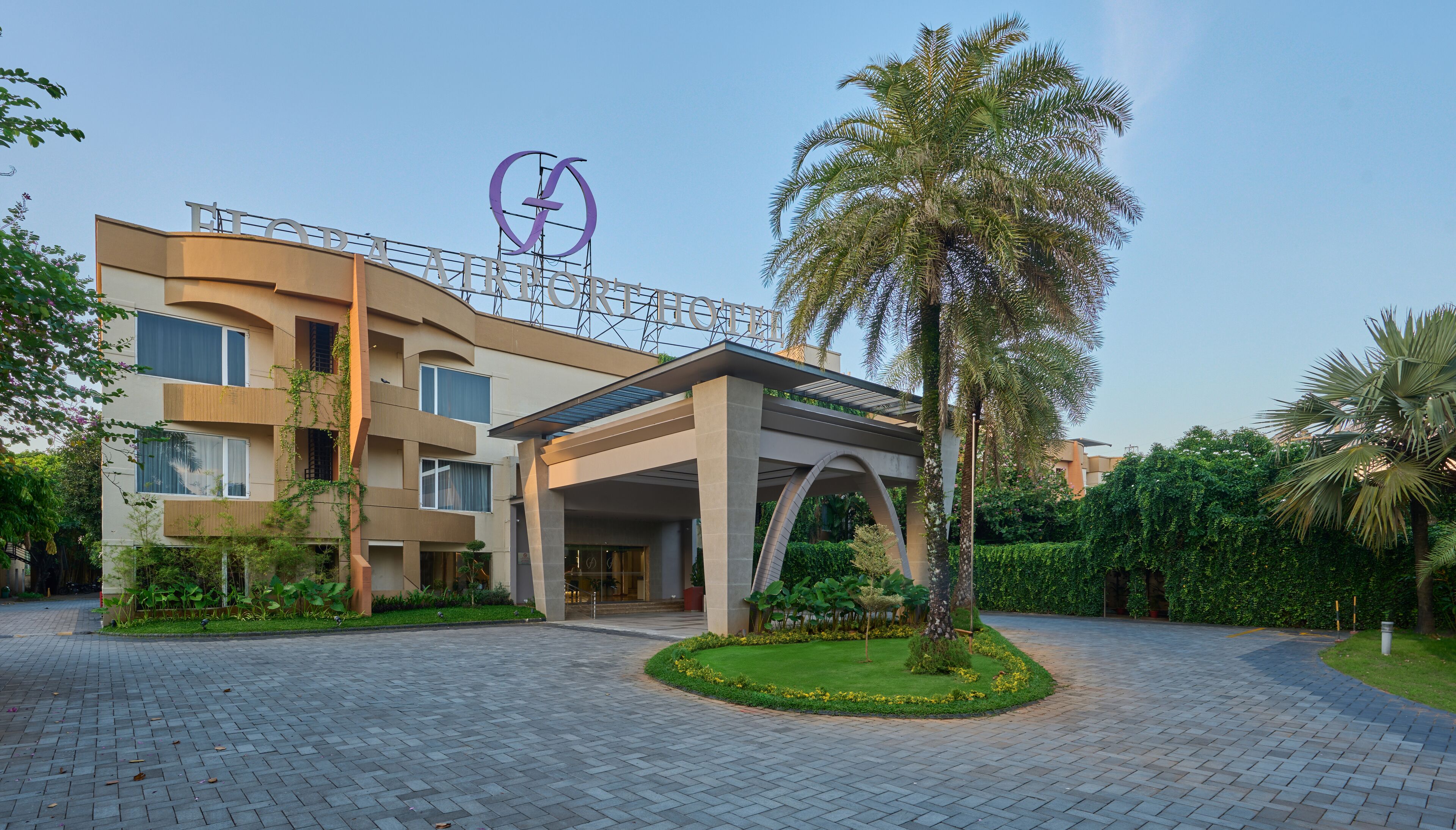 Un hotel modern amb un logotip destacat, un vestíbul cobert i un jardí ben cuidat.