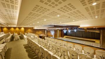 Uma sala de conferências com fileiras de cadeiras, um palco e um teto com padrão quadriculado.