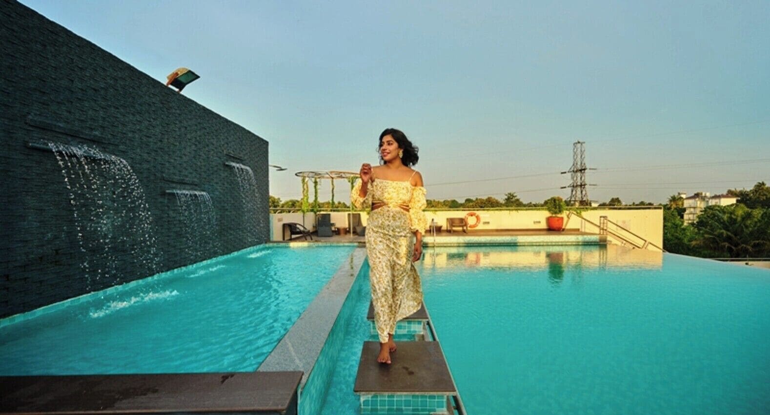 A person standing on a poolside platform with a clear view of a swimming pool and a building with a water feature.