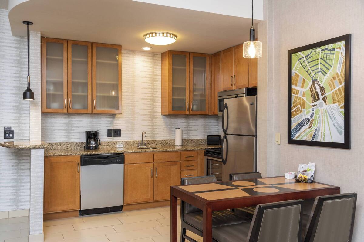 A modern kitchen with wooden cabinets, a stainless steel refrigerator, a coffee maker, and a dining area with a table and chairs.
