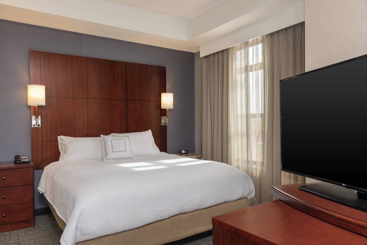 A hotel room with a large bed, two bedside lamps, a flat-screen TV, a wooden nightstand, and a window with curtains.