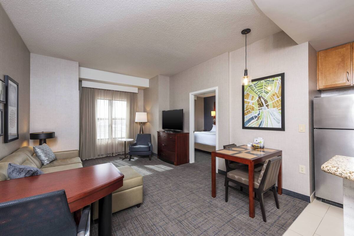 A hotel room with a living area, a dining table, a sofa, a TV, and a kitchen area.