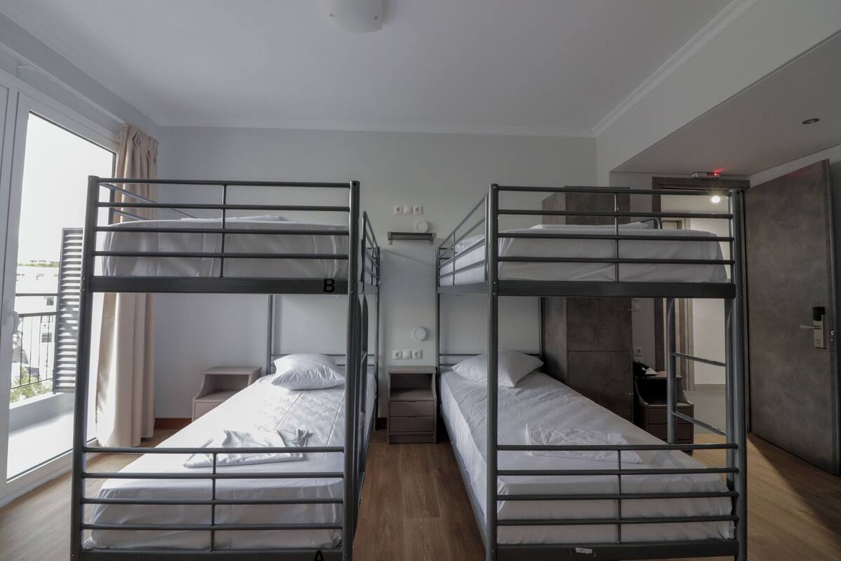 A room with two bunk beds, a small bedside table with a lamp, a window with curtains, and a door with a handle.
