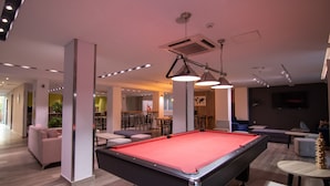 Une table de billard moderne, installée à l’intérieur, avec un éclairage suspendu, entourée d’espaces de détente et d’un bar.