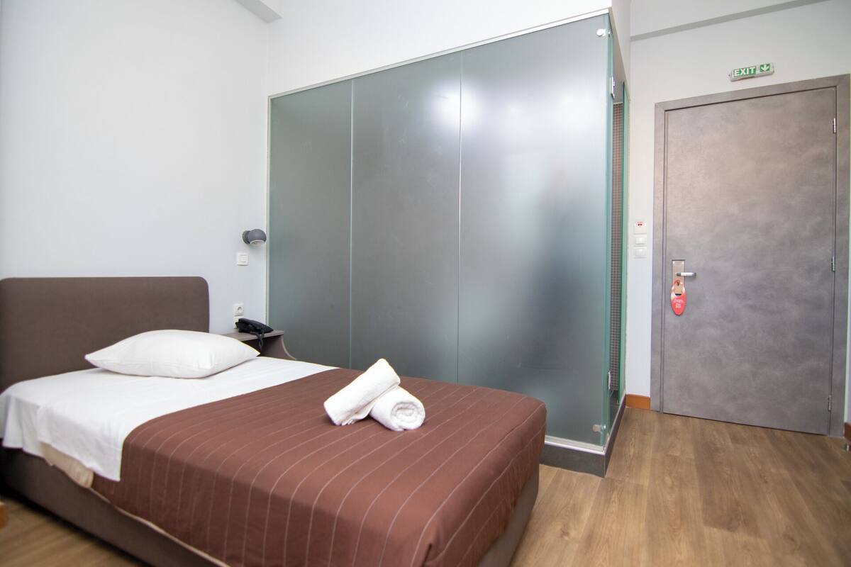 A hotel room with a bed, a nightstand, a telephone, a towel, a door, and an exit sign.
