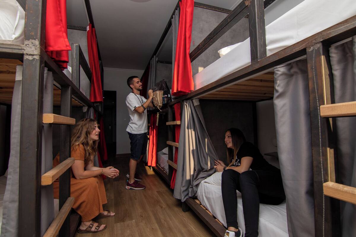 A dormitory room with bunk beds, wooden flooring, and red curtains.