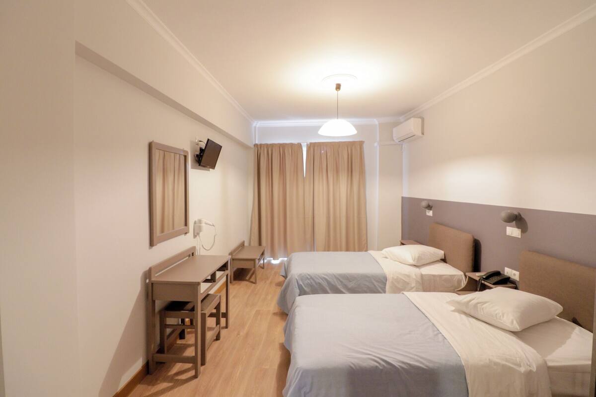 A hotel room with two single beds, a desk with a chair, a TV, and a ceiling light.